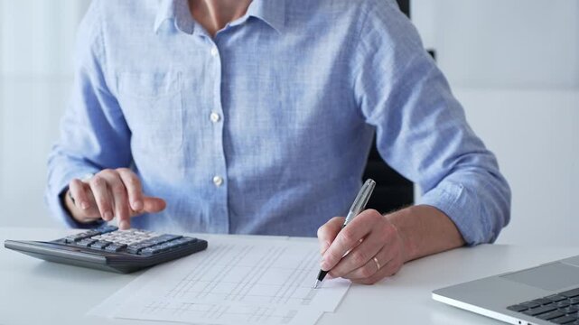 Businessman wearing a blue shirt is using a calculator and taking notes while working on a financial report in the office, close up. Audit and taxes theme in business