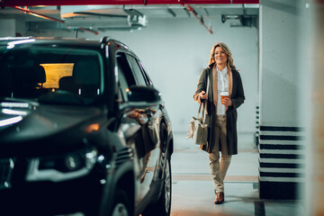 Fototapeta na wymiar Professional businesswoman walking toward her vehicle in a dimly lit underground parking lot, carrying a handbag and takeaway coffee
