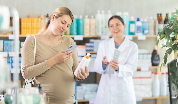 Focused pregnant woman using smartphone to scan barcode on skincare lotion at drugstore, verifying ingredients and safety, while friendly pharmacist providing assistance in background