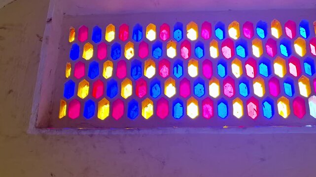 Vibrant stained glass in a honeycomb pattern glows within an arched frame. Sunlight filters through the pink, yellow, and blue panes, casting a colorful radiance inside the Hawa Mahal in Jaipur.