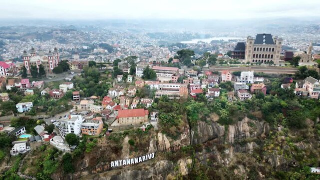 Aerial drone view above Antananarivo with the city skyline spread across surrounding hills, showing dense urban neighborhoods and the large historic palace complex overlooking Madagascar's capital