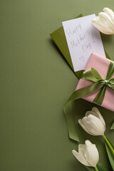 Fototapeta premium Happy Mother's Day handwritten card with pink wrapped gift, green satin ribbon and white tulip arranged on an olive vertical background for a delicate spring celebration of love and appreciation