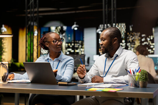 Manager helping young intern in office to complete spreadsheets using laptop, analyzing data. Team leader assisting trainee researching information online on notebook, working overnight