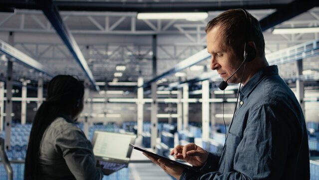 Data center admin talking in audio headset mic, helping clients resolve website hosting problems. Server farm worker on industrial platform helping users keep online services running, camera A