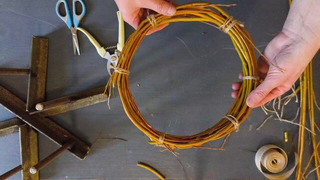 Top down view of hands weaving a rustic wreath base from natural willow branches, authentic DIY home craft hobby and decoration making.
