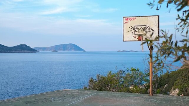Abandoned basketball court with graffiti overlooking the sea. Old, abandoned basketball court with graffiti on the backboard standing on a cliff by the calm blue sea, with distant islands