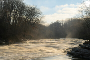 Rushing water flows over Lower Cataract Falls on Mill Creek after heavy rainfall, creating misty cascades through bare winter trees in early morning light, Indiana wilderness waterscape © Douglas