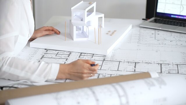 Female architect engrossed in architectural plans and miniature scale model, thoughtfully designing a new building project for modern construction development