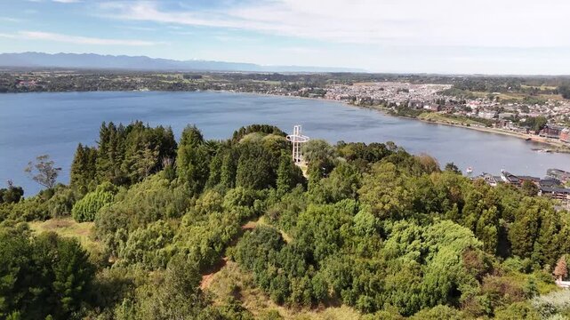 Aerial moves forward toward the iconic illuminated cross on Cerro Philippi, tilting down to reveal the surrounding park trails, dense forest, and Lake Llanquihue in Puerto Varas, Chile