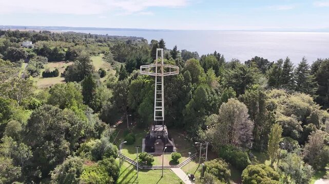 Cinematic aerial pulling back from the monumental white cross on Cerro Philippi to reveal the lush native forest park and the vast blue waters of Lake Llanquihue in Puerto Varas, Chile