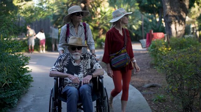 Elderly woman pushes her husband in a wheelchair while their daughter walks beside them in a park in San Diego, California.