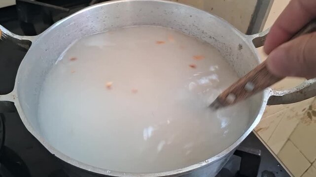 Person's hand continuously stirring creamy white rice or grain porridge in metal pot, cooking footage.