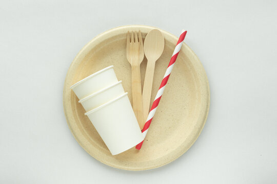 Eco-friendly disposable tableware set with biodegradable plate, wooden fork and spoon, paper cups, and striped paper straw on minimal background.