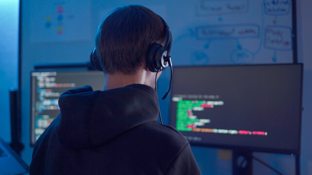 Software developer wearing headset reviewing program code on monitor in low light workspace