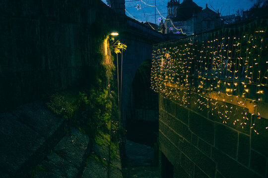 Atmospheric night view of a narrow passage by the old stone bridge in Amarante, Portugal, featuring mossy walls and warm Christmas fairy lights glowing against the dark twilight.