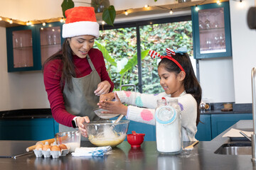Asian mother and daughter in Santa hat, striped headband mixing batter in home kitchen with sifter