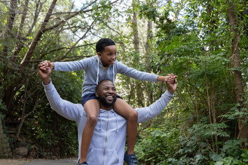 Naklejka premium African American father and son walking wooded path, carrying son on shoulders in gray hoodies