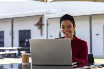 Fototapeta premium African American woman wearing burgundy top working on silver laptop at patio table with coffee cup