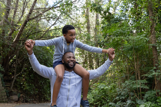 African American father and son walking wooded path, carrying son on shoulders in gray hoodies