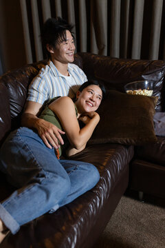 Couple cuddling on leather sofa in living room, woman reclining on cushion, popcorn bowl resting
