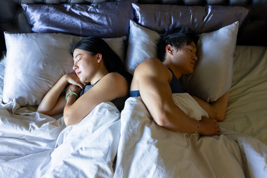 Asian couple lying back-to-back on king bed in bedroom wearing dark sleeveless tops, bracelets