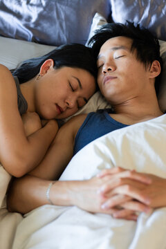 Asian couple sleeping in bed under white duvet with gray pillows and clasped hands