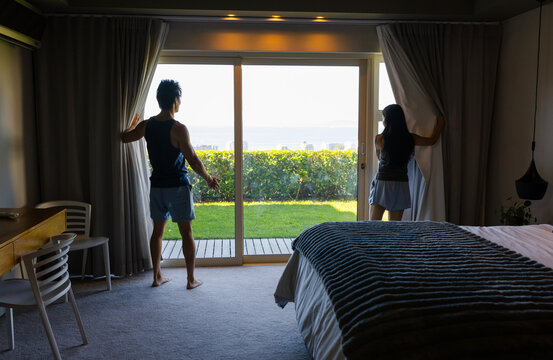 Asian couple standing at hotel bedroom sliding glass door pulling back heavy curtains revealing sea