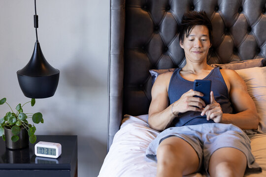 Asian male reclining on bed at home, holding smartphone near tufted headboard, clock and plant