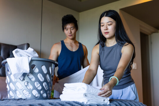 Asian couple in tank tops folding white towels on bed, woman stacking towels into laundry basket