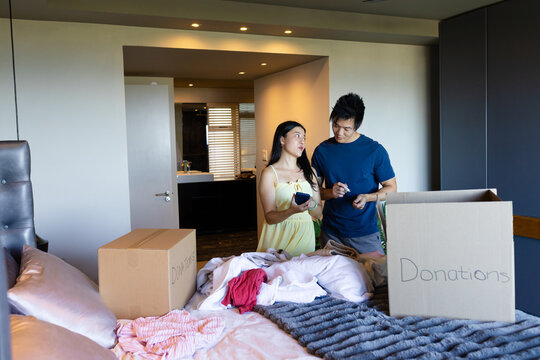 Asian couple standing and sorting donations at bedroom foot of bed with boxes, smartphone