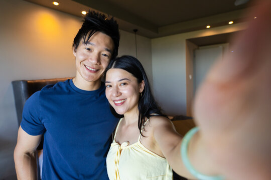 Smiling couple wearing blue shirt posing in hotel room, holding phone selfie, wristband, headboard