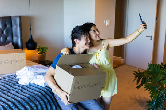Asian couple packing Donations boxes and taking selfie with smartphone in bedroom by striped bed