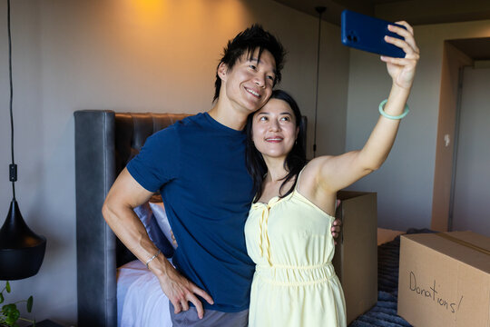 Asian couple in casual wear posing, taking selfie in bedroom with blue phone and Donations boxes