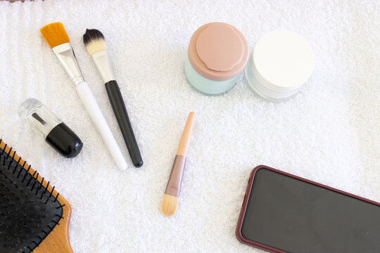 Makeup brushes, cream jars, clear bottle are resting on white terry towel on vanity, pastel accents