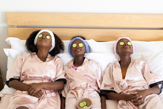 Reclining mother with daughters wearing pink satin robes and headbands on bed with cucumber slices