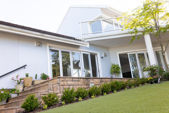Modern two-story house facade showcasing stone wall, sliding glass doors, potted plants under sun