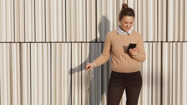 A cheerful woman in a beige sweater stands with her suitcase, checking a travel app on her smartphone against a modern, ribbed architectural background.