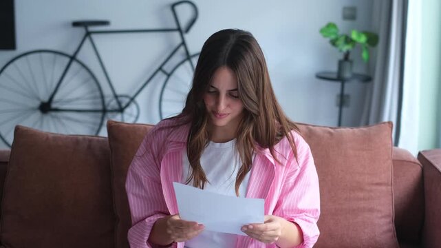 Smiling attractive young woman indoor sit on sofa at home reading letter. Pleased female holds in hands sheet pleased by good news written in official document. Bank notice, happy client concept

