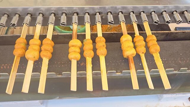 Row automatically rotating meatballs on nine skewers roasting above charcoal barbecue, Thai traditional street food