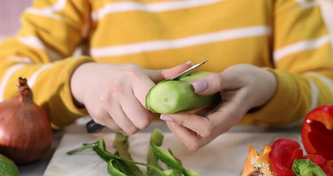 Woman peeling fresh cucumber at grey table with vegetables indoors, closeup. Camera moving forward