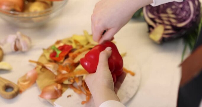 Woman removing core of fresh bell pepper with knife at light table with leftovers and vegetables indoors, closeup. Camera moving forward