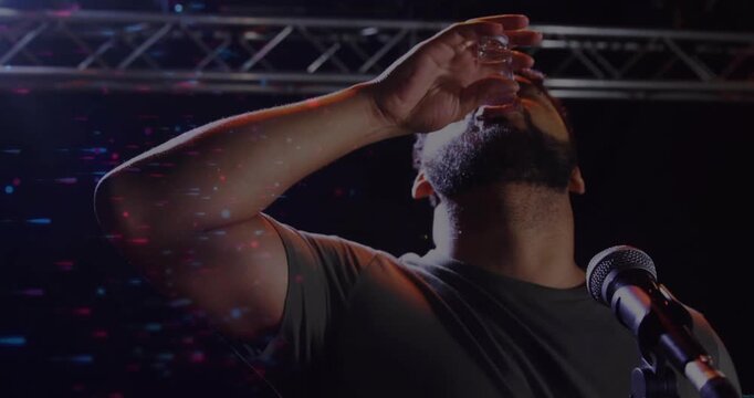 Host lifting shot glass starting drinking on comedy stage raising arm cheering amid light specks