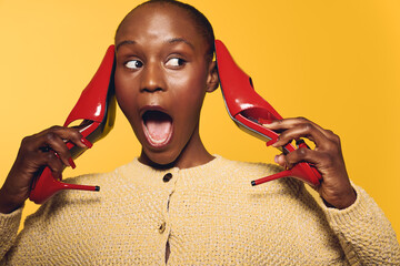 Surprised black woman with dark skin holds red high heels near ears, wearing beige sweater. Bright...
