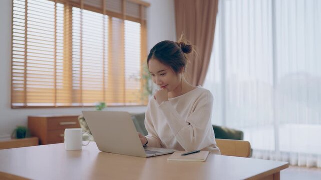 Young Asian woman using laptop computer for business studying, having video call with online virtual webinar training meeting, writing notes. Focused student female working in living room at home