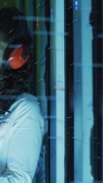 Vertical video: Woman tech monitoring rack in muffs, LEDs flashing and initiating AR overlays