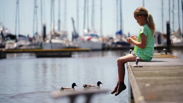 A young blonde girl in a green outfit sits on a wooden dock, dangling her feet. She throws food to ducks swimming in the marina, with sailboats blurred in the background on a sunny day.