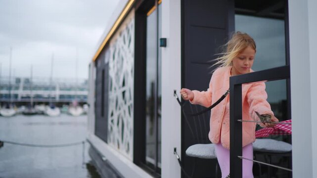 A young blonde girl in a pink jacket plays on the deck of a modern houseboat. Her hair blows in the wind as she stands by the water in a marina on a cloudy day