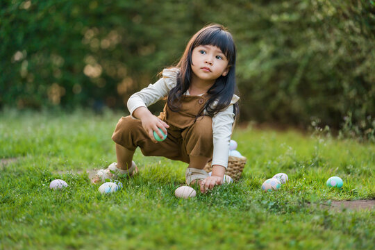 child girl playing and hunting for Easter eggs into a basket on green grass in garden