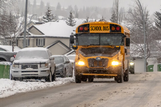 Calgary, Alberta, Canada. March 16, 2026. A yellow school bus driving through a snowy residential street during a winter storm.