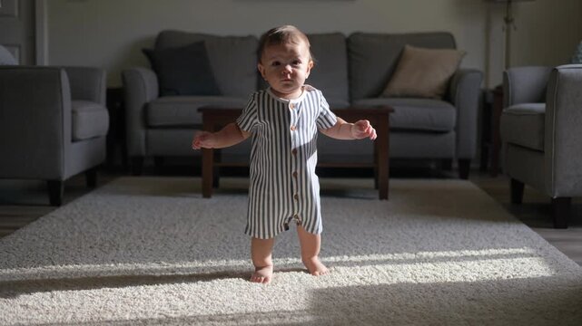 A young toddler wearing a blue and white striped romper takes brave first steps across a sunlit living room carpet toward the camera.